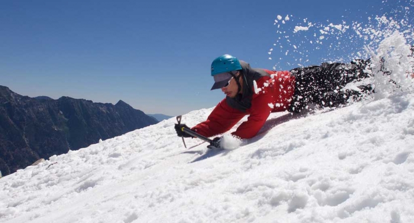 A person wearing mountaineering gear slides down a snowy incline on their belly.